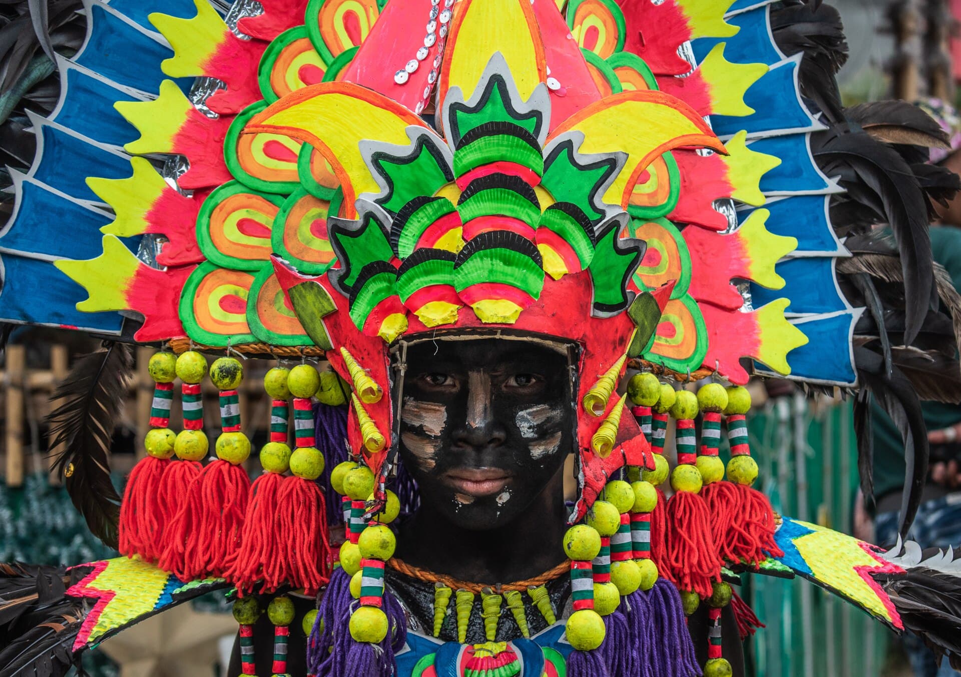 Wide shot of a Kalibo tribe in soot and feathered headdresses dancing the sadsad down a packed street on the final Sunday