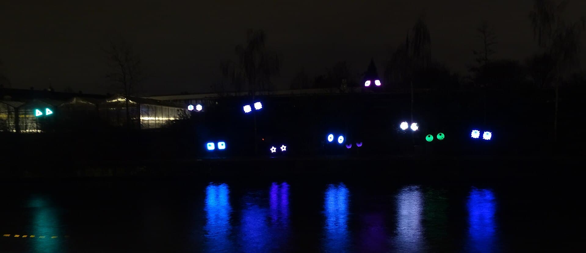 Wide shot of a lit artwork reflected in an Amsterdam canal at dusk, with canal houses on both banks