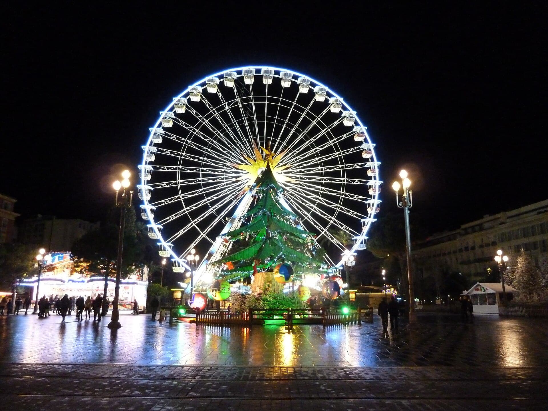 Wide shot of an illuminated carnival float on Place Masséna at night in Nice with crowds in the grandstands
