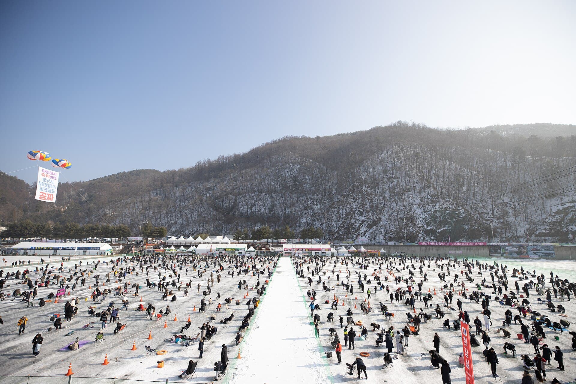 Wide aerial view of the frozen Hwacheoncheon Stream dotted with ice fishing holes and crowds at the Hwacheon Sancheoneo Ice Festival