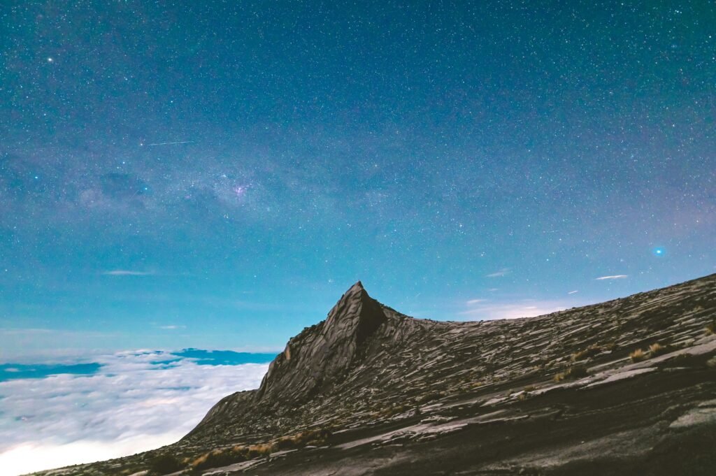 Summit of Mount Kinabalu under a starry night sky with visible Milky Way, overlooking a sea of clouds and rugged rocky terrain