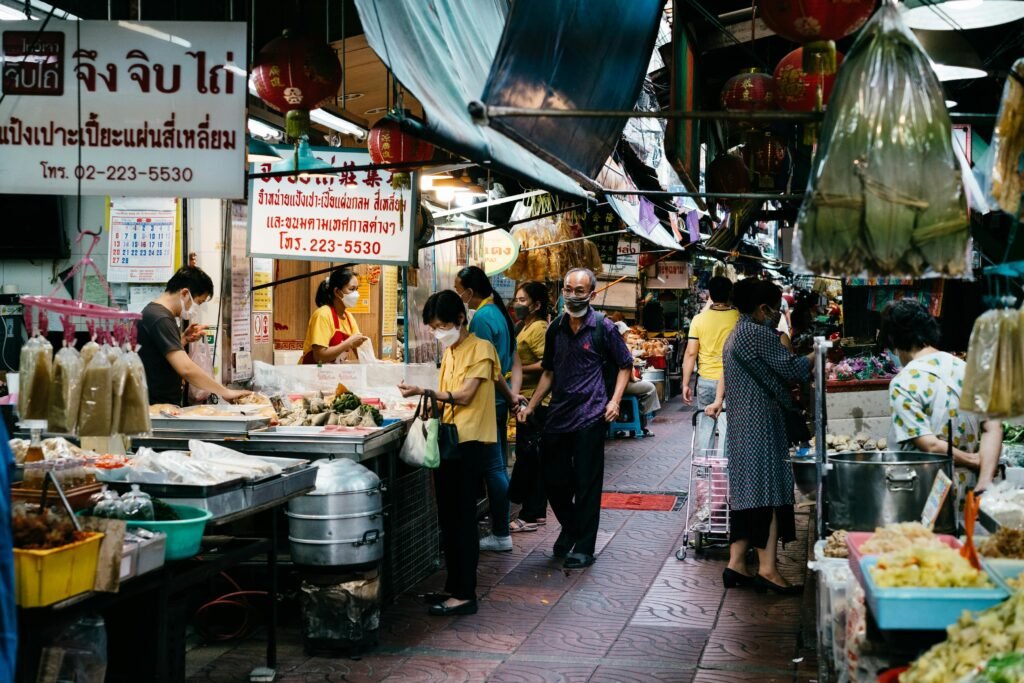 Busy local food market in Bangkok, Thailand, with vendors and shoppers wearing masks, surrounded by street food stalls and colorful signage